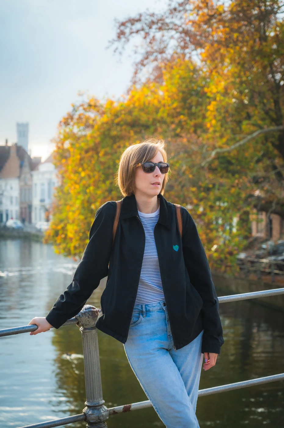 AWoman with sunglasses leaning on a railing beside a canal, autumn trees in the background.